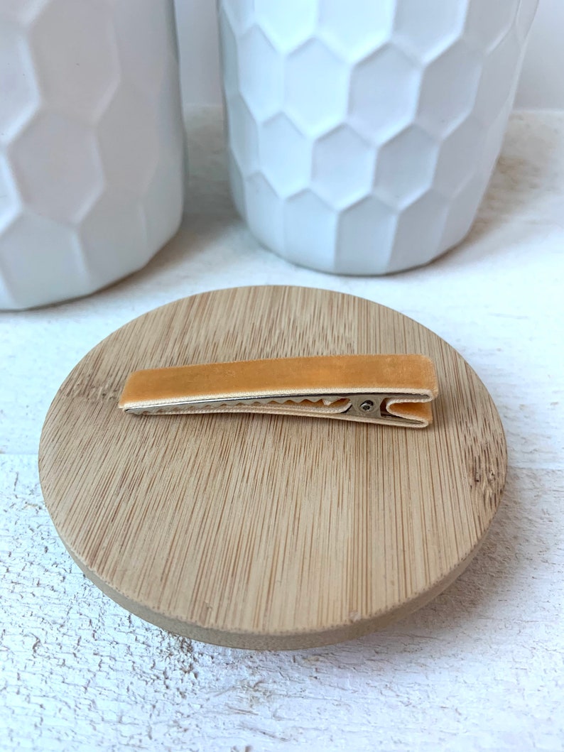 May include: A light brown velvet hair clip with a gold metal clasp. The clip is sitting on a wooden surface.