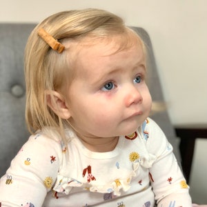 Puede incluir: Una ni&ntilde;a peque&ntilde;a con cabello rubio que lleva una camisa blanca de manga larga con un estampado colorido de animales y flores. La camisa tiene un cuello con volantes. La ni&ntilde;a lleva una pinza para el pelo de madera en el pelo.