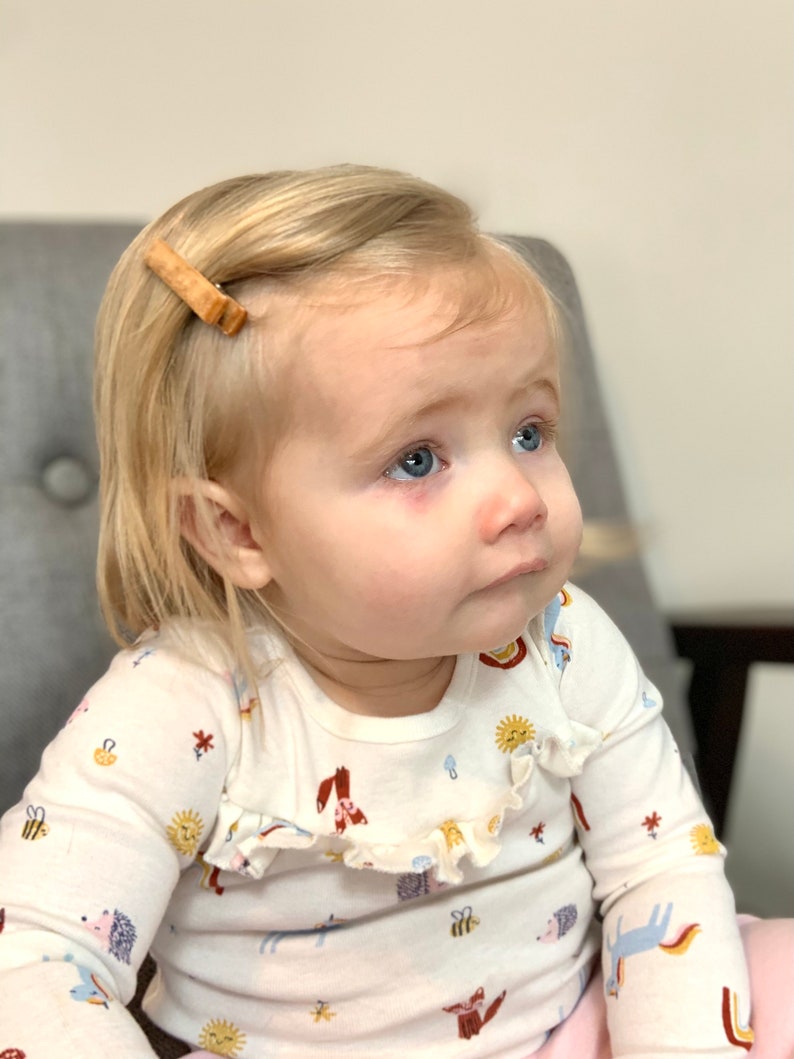 Puede incluir: Una ni&ntilde;a peque&ntilde;a con cabello rubio que lleva una camisa blanca de manga larga con un estampado colorido de animales y flores. La camisa tiene un cuello con volantes. La ni&ntilde;a lleva una pinza para el pelo de madera en el pelo.