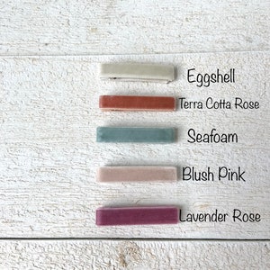 Puede incluir: Cinco pinzas para el pelo de terciopelo en diferentes colores: c&aacute;scara de huevo, rosa terracota, espuma de mar, rosa rubor y rosa lavanda. Las pinzas est&aacute;n dispuestas verticalmente sobre una superficie de madera blanca.