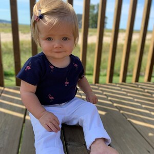 May include: A young child wearing a navy blue shirt with pink flamingo print and white pants sits on a wooden deck.