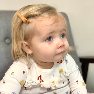 May include: A young child with blonde hair wearing a white long-sleeved shirt with a colorful pattern of animals and flowers. The shirt has a ruffled neckline. The child is wearing a wooden hair clip in their hair.