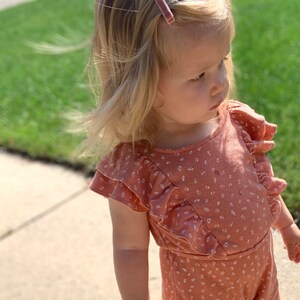 Puede incluir: Una ni&ntilde;a peque&ntilde;a con un mono rosa de flores con mangas con volantes y una pinza para el pelo rosa.