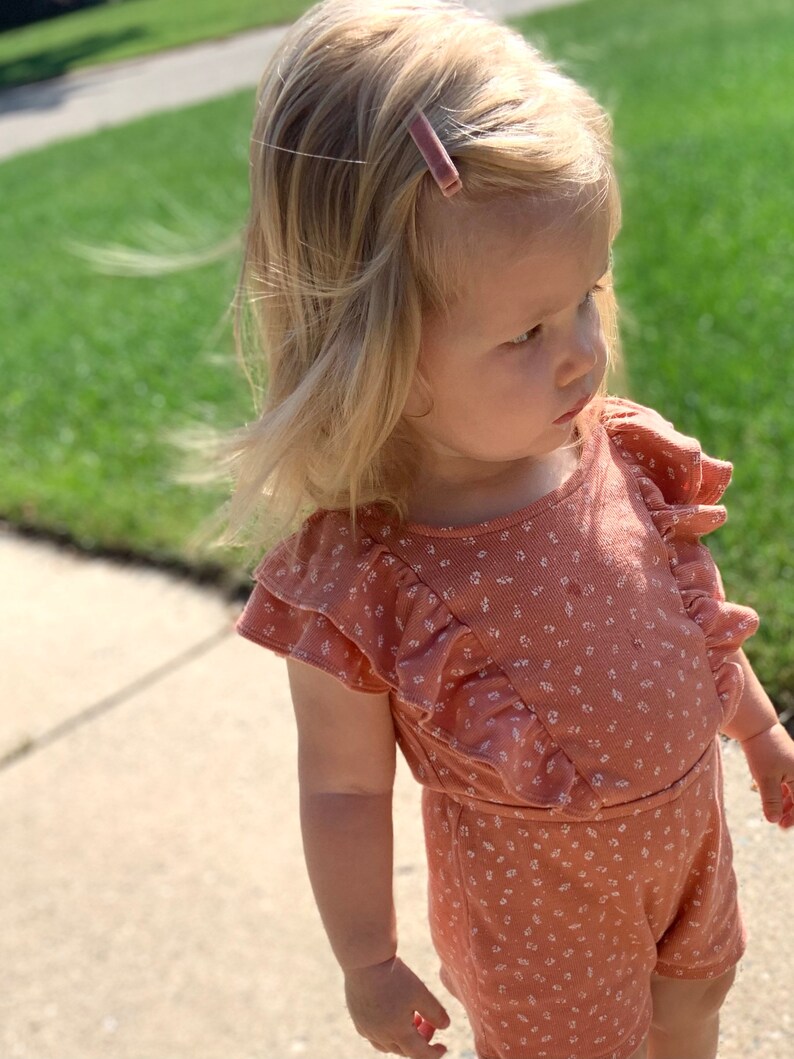 Puede incluir: Una ni&ntilde;a peque&ntilde;a con un mono rosa de flores con mangas con volantes y una pinza para el pelo rosa.
