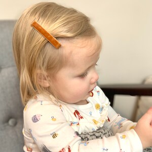 May include: A young child with blonde hair wearing a white long-sleeved shirt with a colorful animal print. The child is wearing a brown velvet barrette in their hair.