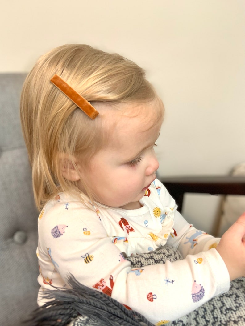 May include: A young child with blonde hair wearing a white long-sleeved shirt with a colorful animal print. The child is wearing a brown velvet barrette in their hair.