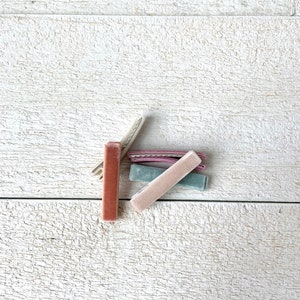 Puede incluir: Cuatro pinzas para el pelo de terciopelo en diferentes colores: rosa polvoriento, rosa claro, azul claro y marr&oacute;n oscuro. Las pinzas est&aacute;n dispuestas en una pila sobre una superficie de madera blanca.