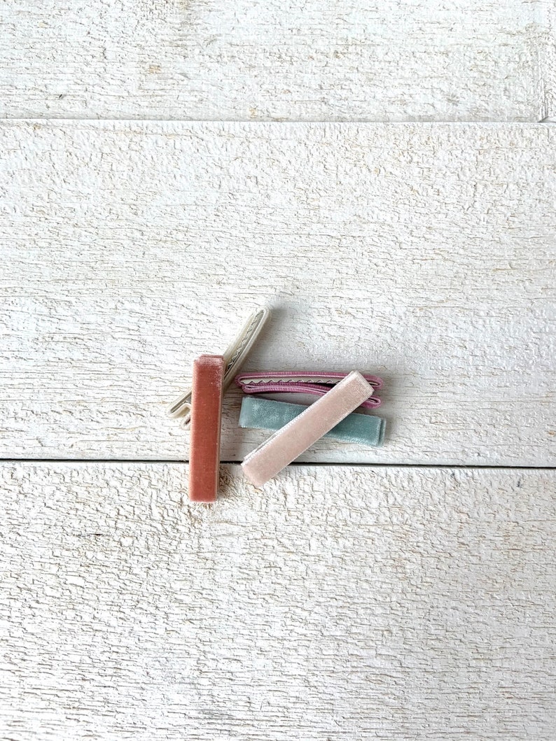 Puede incluir: Cuatro pinzas para el pelo de terciopelo en diferentes colores: rosa polvoriento, rosa claro, azul claro y marr&oacute;n oscuro. Las pinzas est&aacute;n dispuestas en una pila sobre una superficie de madera blanca.