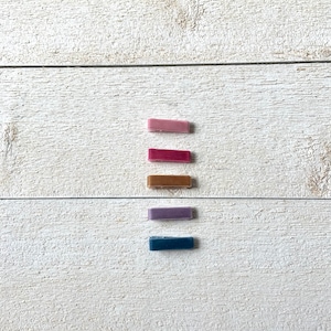 May include: Five velvet hair clips in different colors: pink, burgundy, brown, purple, and blue. The clips are arranged vertically on a white wooden surface.