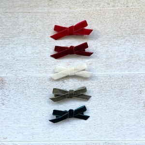 May include: Five velvet hair bows in different colors: red, white, gray, dark green, and black. The bows are arranged vertically on a white wooden background.