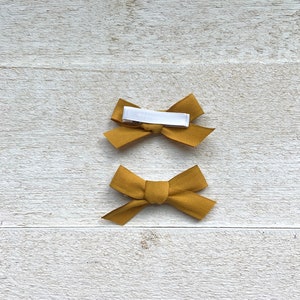 May include: Two mustard yellow fabric hair bows with white alligator clips. The bows are tied in a simple knot.