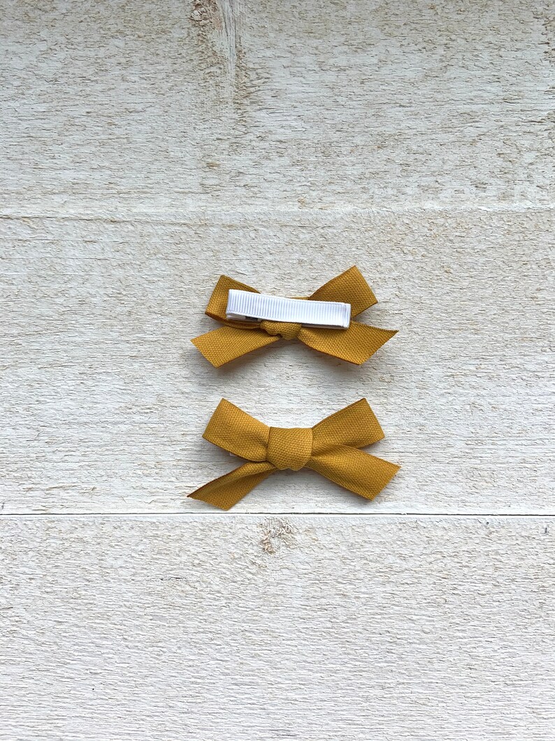 May include: Two mustard yellow fabric hair bows with white alligator clips. The bows are tied in a simple knot.