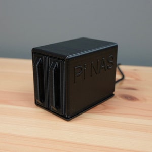 K&ouml;nnte beinhalten: Ein schwarzes, 3D-gedrucktes Geh&auml;use mit dem Text "Pi NAS" auf der Vorderseite. Das Geh&auml;use hat zwei Steckpl&auml;tze f&uuml;r Festplatten.