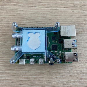 May include: A Raspberry Pi computer board with a clear plastic heatsink attached. The board has a blue and green circuit board with several ports, including HDMI, USB, and Ethernet. The board is mounted on a clear plastic frame with screws.