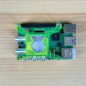 Puede incluir: Una placa de computadora Raspberry Pi 5 verde con una carcasa de plástico transparente y un disipador de calor plateado. La placa tiene una etiqueta que dice "Raspberry Pi 5" y "HAT GPIO Interface".