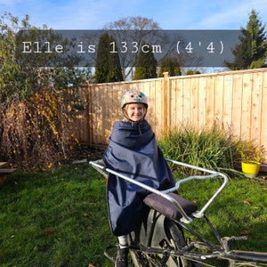 May include: A young person wearing a colourful helmet sits in a blue rain cover on a silver bike trailer. The text "Elle is 133cm (4'4)" is visible in the image.