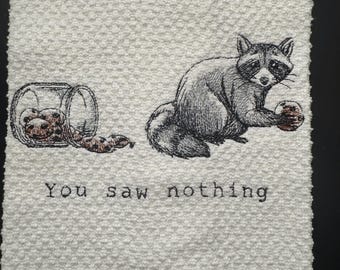 Könnte beinhalten: Weißes Küchentuch mit einer Schwarz-Weiß-Illustration eines Waschbären. Der Waschbär hält einen Keks, mit einem verschütteten Glas Kekse in der Nähe. Der Text "You saw nothing" ist auf das Handtuch gestickt.