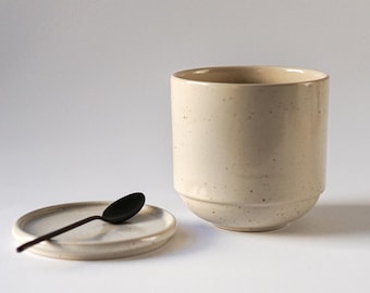 May include: A white ceramic jar with a speckled finish and a matching lid. The jar has a slightly rounded shape and a wide opening. A small black spoon is resting on the lid.