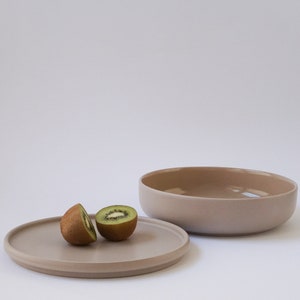 Op de afbeelding: Twee helften van een kiwi op een beige keramisch bord naast een grotere beige keramische kom.