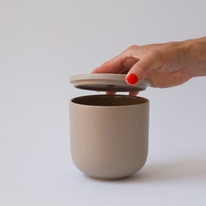 Può includere: Un vaso in ceramica beige con coperchio abbinato. Il vaso ha una base arrotondata e un corpo cilindrico. Il coperchio viene sollevato, rivelando l'interno. Il vaso viene probabilmente utilizzato per la conservazione. Lo sfondo è bianco.