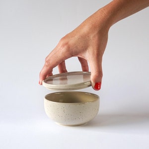 Könnte beinhalten: Eine beige Keramik Schüssel mit gesprenkeltem Finish und einem passenden Deckel. Der Deckel wird von einer Hand von der Schüssel abgehoben.