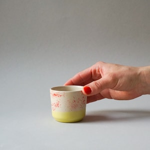 Peut inclure: Une petite tasse en céramique jaune avec un bord blanc et des taches rouges. La tasse est posée sur une surface grise.