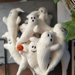 Könnte beinhalten: Eine Gruppe weißer Filz-Geister-Ornamente mit schwarzen Augen und Mündern. Die Geister sind in einem Stapel angeordnet, wobei ein Geist einen kleinen orangefarbenen Ball hält.
