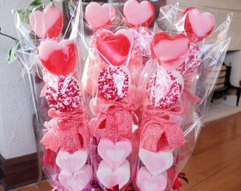 Può includere: Un bouquet di spiedini di caramelle, ciascuno avvolto in cellophane trasparente. Gli spiedini presentano lecca-lecca a forma di cuore, dischi ricoperti di caramelle e marshmallow a forma di cuore in tonalità rosse e rosa. La base è decorata con nastri rosa e rossi.