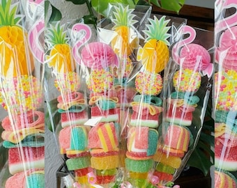 Può includere: Un bouquet di spiedini di caramelle colorate con topper a forma di fenicottero rosa e dettagli di ananas. Gli spiedini sono avvolti in cellophane trasparente e disposti in un cestino verde e bianco.