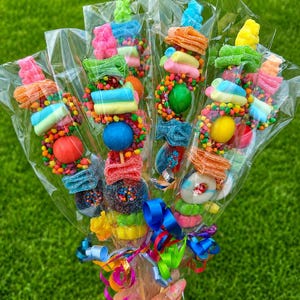 May include: A bouquet of four candy skewers wrapped in clear plastic, each featuring a variety of colorful gummy candies, marshmallows, and round sugar-coated sweets. The skewers are tied together at the base with bright curling ribbons.