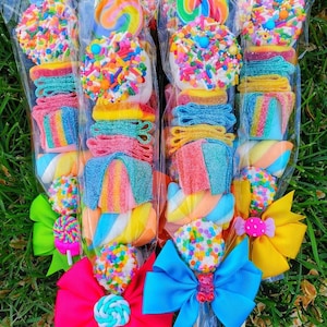 May include: Candy skewers featuring a variety of colorful treats, including lollipops, gummy candies, marshmallows, and sprinkles. Each skewer is wrapped in clear cellophane and adorned with a decorative bow. The skewers are arranged on a bed of green grass.