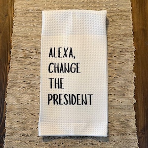 May include: White kitchen towel with black embroidered text that reads "Alexa, change the president."