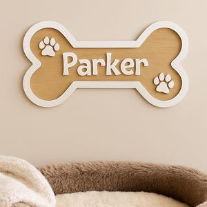Peut inclure: Panneau en bois en forme d'os de chien avec le nom "Parker" en blanc, orné d'empreintes de pattes. En dessous, un lit pour chien marron avec un intérieur moelleux de couleur claire et une couverture crème.