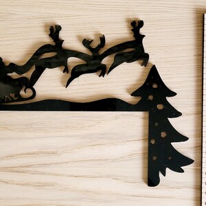 May include: Black metal silhouette of Santa Claus in a sleigh pulled by reindeer flying over a snowdrift and a Christmas tree. The silhouette is approximately 17 inches tall.