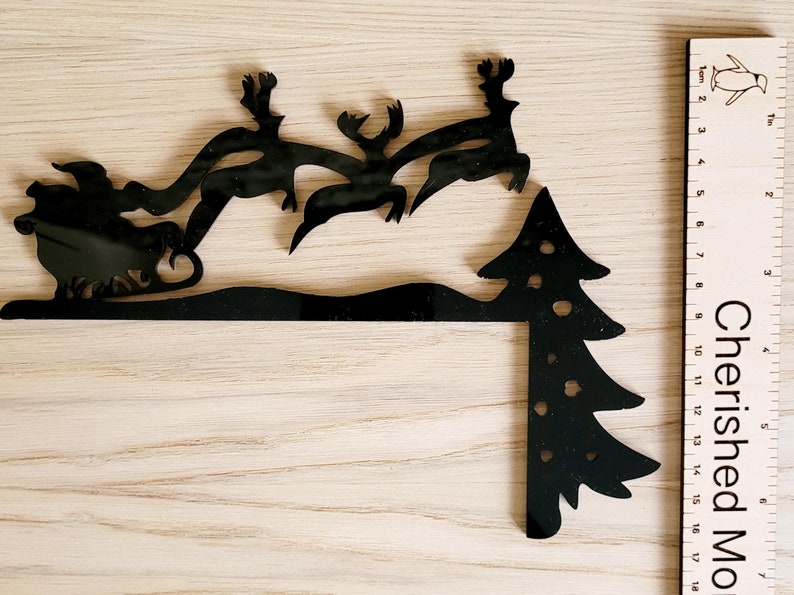 May include: Black metal silhouette of Santa Claus in a sleigh pulled by reindeer flying over a snowdrift and a Christmas tree. The silhouette is approximately 17 inches tall.