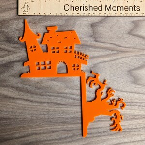 May include: Orange Halloween-themed cutout of a haunted house with a spooky tree. The house has a pointed roof, windows, and a door. The tree has branches with spooky details.