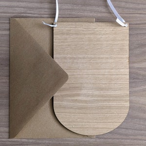 K&ouml;nnte beinhalten: Ein brauner Papierumschlag mit einem wei&szlig;en Band darum gebunden, und ein Holzschild mit abgerundeter Oberseite und einem wei&szlig;en Band darum gebunden.