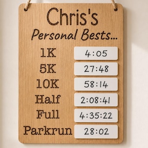 May include: A rectangular light wood plaque with a rope hanger featuring the engraved text Chris's Personal Bests... and a list of running distances including 1K, 5K, 10K, Half, Full, and Parkrun, paired with white rectangular inserts showing times.