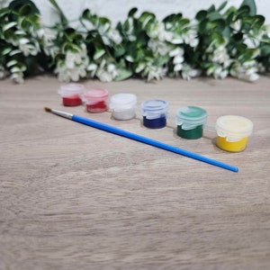 K&ouml;nnte beinhalten: Sechs kleine Farbt&ouml;pfe mit Deckeln in Rot, Rosa, Wei&szlig;, Blau, Gr&uuml;n und Gelb. Ein blauer Pinsel liegt auf einer Holzoberfl&auml;che.