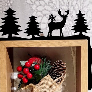 May include: Black metal shelf decor with a silhouette of a deer with a snowflake on its antlers, two pine trees, and a snow-covered ground. The decor is mounted on a wooden shelf with a burlap-wrapped bouquet of pine needles, red berries, and a pine cone.