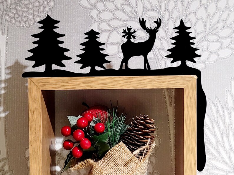 May include: Black metal shelf decor with a silhouette of a deer with a snowflake on its antlers, two pine trees, and a snow-covered ground. The decor is mounted on a wooden shelf with a burlap-wrapped bouquet of pine needles, red berries, and a pine cone.