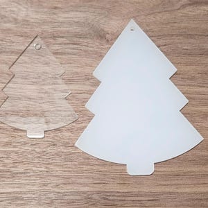 May include: Two white acrylic Christmas tree decorations, one with a hole at the top and the other with a hole at the top and a hole in the centre. The decorations are on a wooden surface.