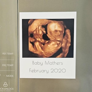 May include: A printed photograph of a baby ultrasound scan, showing a detailed image of a developing fetus. The photo is labeled "Baby Mathers February 2020" in a simple, elegant font. The photo is attached to a refrigerator.