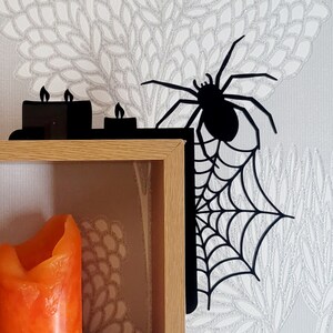 May include: Black metal shelf bracket with a spider and web design. The bracket is mounted on a wooden shelf with a lit orange candle inside.