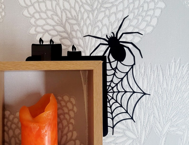 May include: Black metal shelf bracket with a spider and web design. The bracket is mounted on a wooden shelf with a lit orange candle inside.