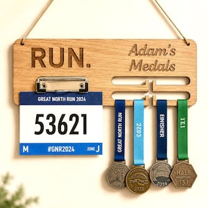 May include: A wooden medal display board with the word "RUN" and "Adam's Medals" engraved. It holds a race bib with the number 53621 and four medals with ribbons. The medals are for the Great North Run and a half marathon.