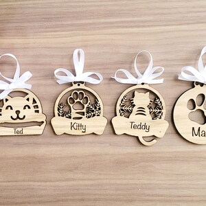 May include: Four wooden Christmas decorations with personalised names. Each decoration features a different design: a cat in a bed, a paw print, a cat, and a paw print. The decorations are decorated with white ribbon.