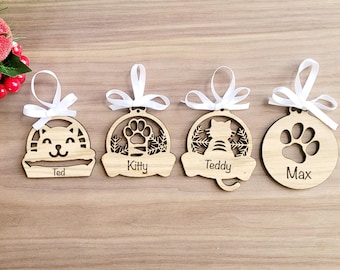Puede incluir: Cuatro adornos navideños de madera con nombres personalizados. Cada adorno presenta un diseño diferente: un gato en una cama, una huella de pata, un gato y una huella de pata. Los adornos están decorados con una cinta blanca.
