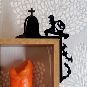 May include: Black metal shelf bracket shaped like a spooky Halloween scene with a tombstone and a ghoul. The bracket is mounted on a wooden shelf with an orange candle inside.