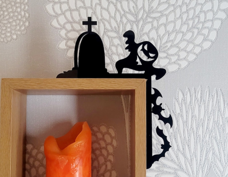 May include: Black metal shelf bracket shaped like a spooky Halloween scene with a tombstone and a ghoul. The bracket is mounted on a wooden shelf with an orange candle inside.